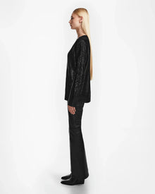 A woman with long straight blonde hair stands in profile, wearing the Rudsak ELLE - 8524597 Black V-neck sweater, black pants, and boots, set against a plain white background.