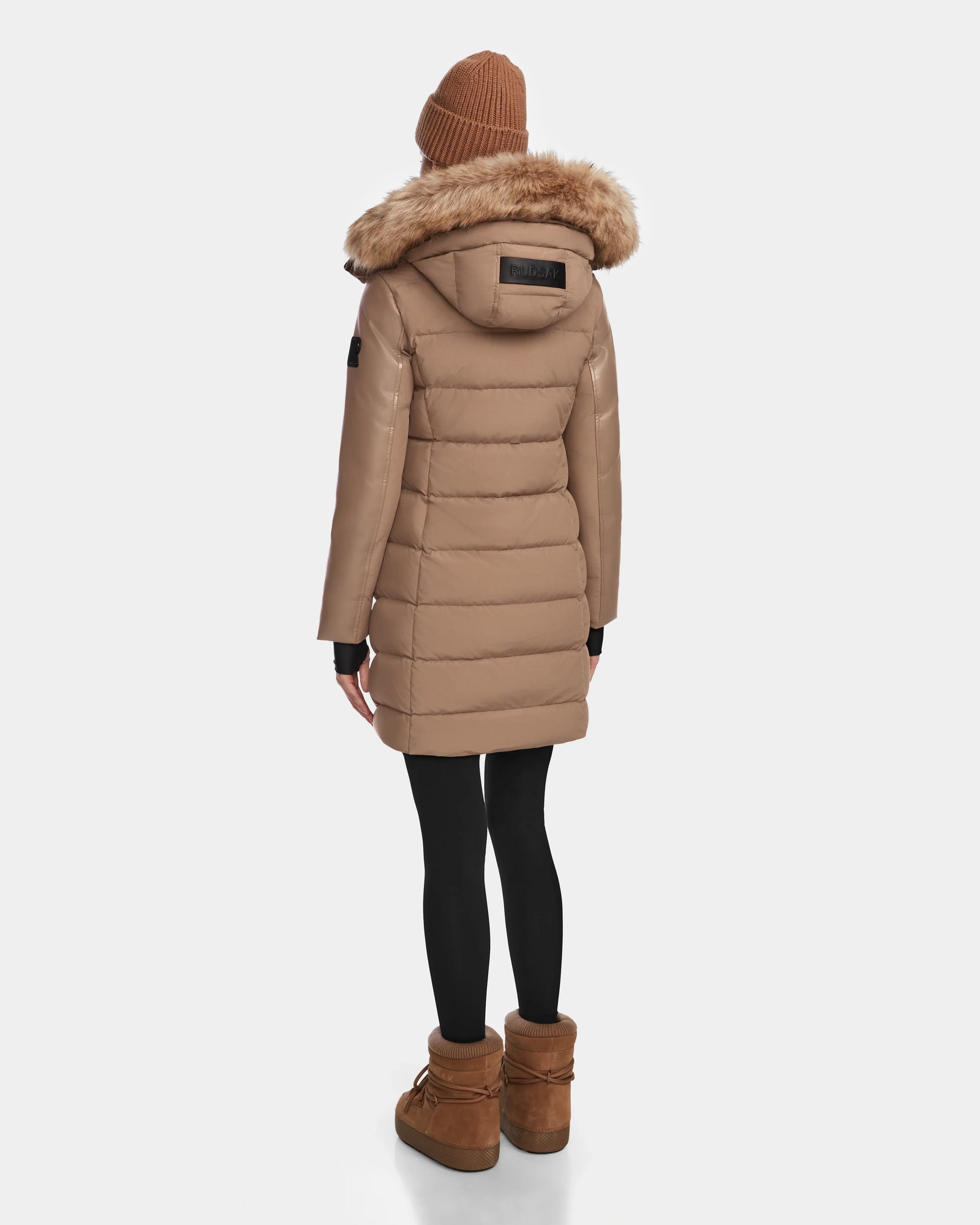 A person wears the Rudsak ELSA - L123535 Sand women’s quilted down puffer with a fur-trimmed hood, brown beanie, black leggings, and brown boots, facing away against a white background. The coat features RDS-certified down for sustainability.