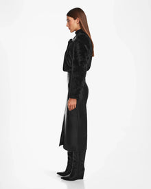 A woman stands in profile against a white background, wearing Rudsak’s EVELYN - 8124791 Black faux fur jacket, paired with a long black leather skirt featuring a front slit and sleek black knee-high boots. Her straight brown hair is loose.