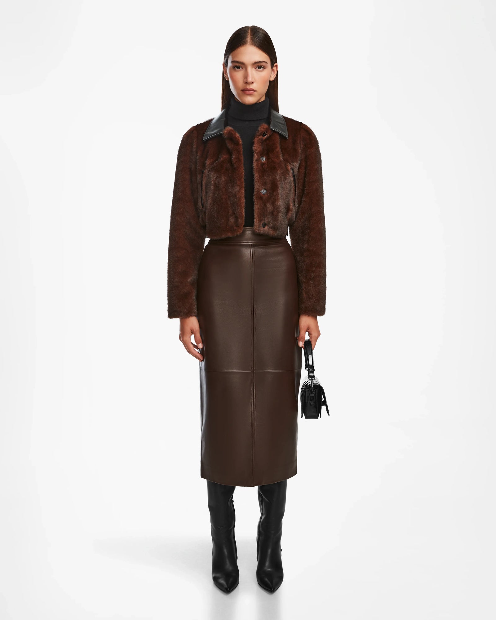 A woman faces forward against a white background, wearing the Rudsak EVELYN - 8124791 Espresso faux fur jacket, styled with a dark brown leather midi skirt, black turtleneck, knee-high boots, and a small black handbag.