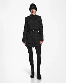 A person wearing the Rudsak FRIDA M - L124524 Black women’s mid-length down jacket with a belt, black tights, boots, and a knit beanie stands facing forward against a plain white background.