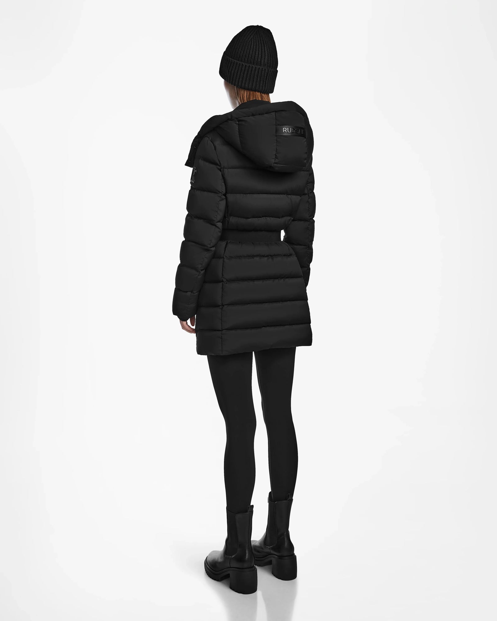 A person wears the Rudsak FRIDA M - L124524 Black womens mid-length down jacket, paired with black leggings, boots, and a knit beanie, standing facing away against a plain white background.