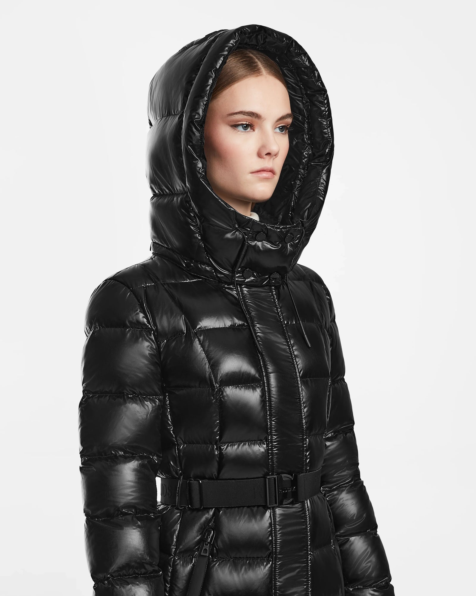 A woman in the Rudsak FRIDA S - L124526 Black shiny mid-length down jacket with hood stands against a plain white background, looking to the side.