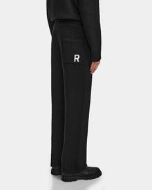 Someone wearing the Rudsak GEIGER - 9524512 Black M relaxed fit lounge pants with a patch pocket featuring a white R stands against a plain light background, paired with black shoes and a black top.