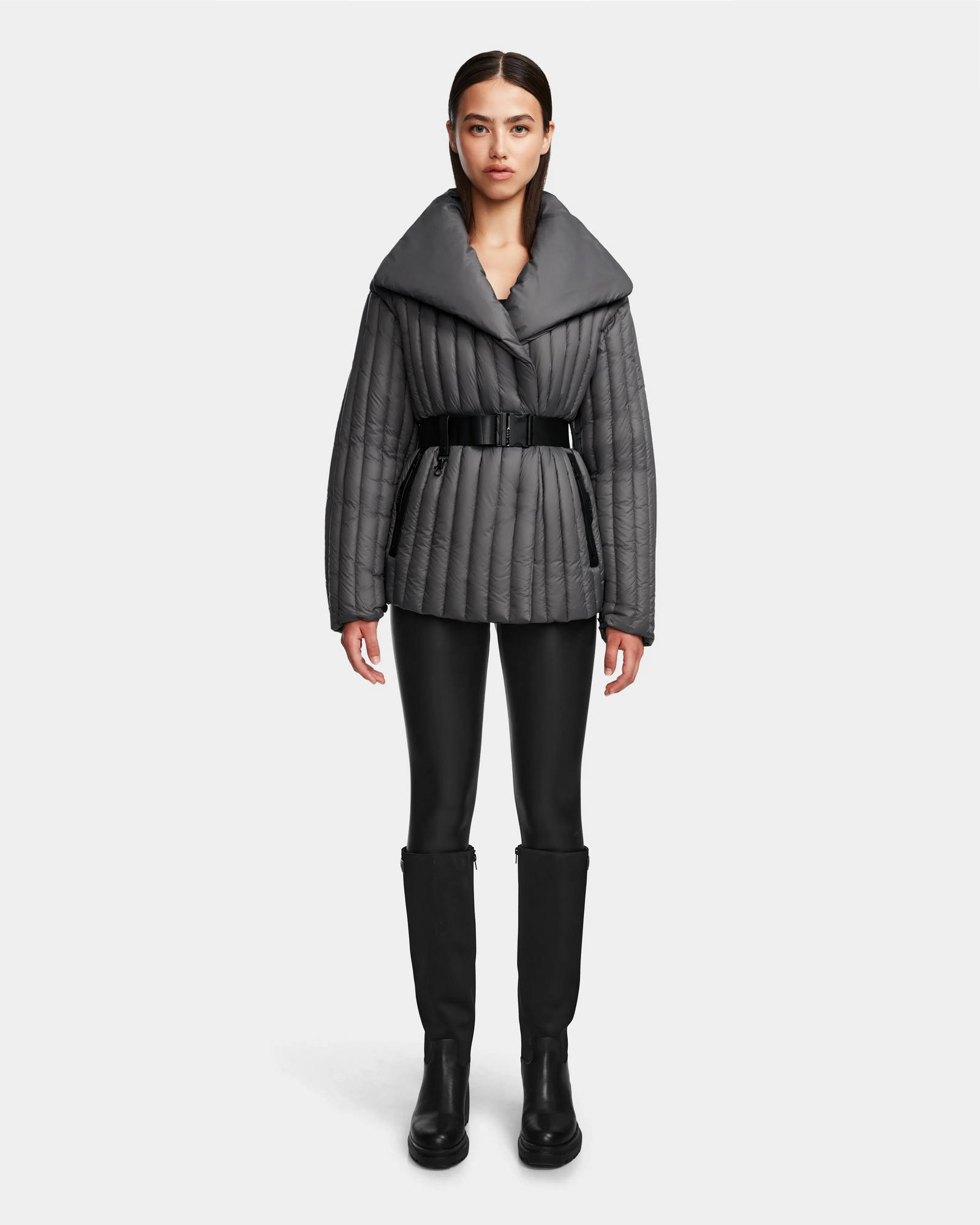 A woman faces forward on a plain white background, wearing the Rudsak INES T - 8124625 Grey quilted jacket with a wide collar, black belt, black leggings, and knee-high black boots.