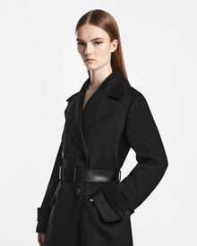 A woman with straight, light brown hair showcases the Rudsak IVE - L124560 Black double-breasted coat with a black belt, looking confidently at the camera against a plain white background.