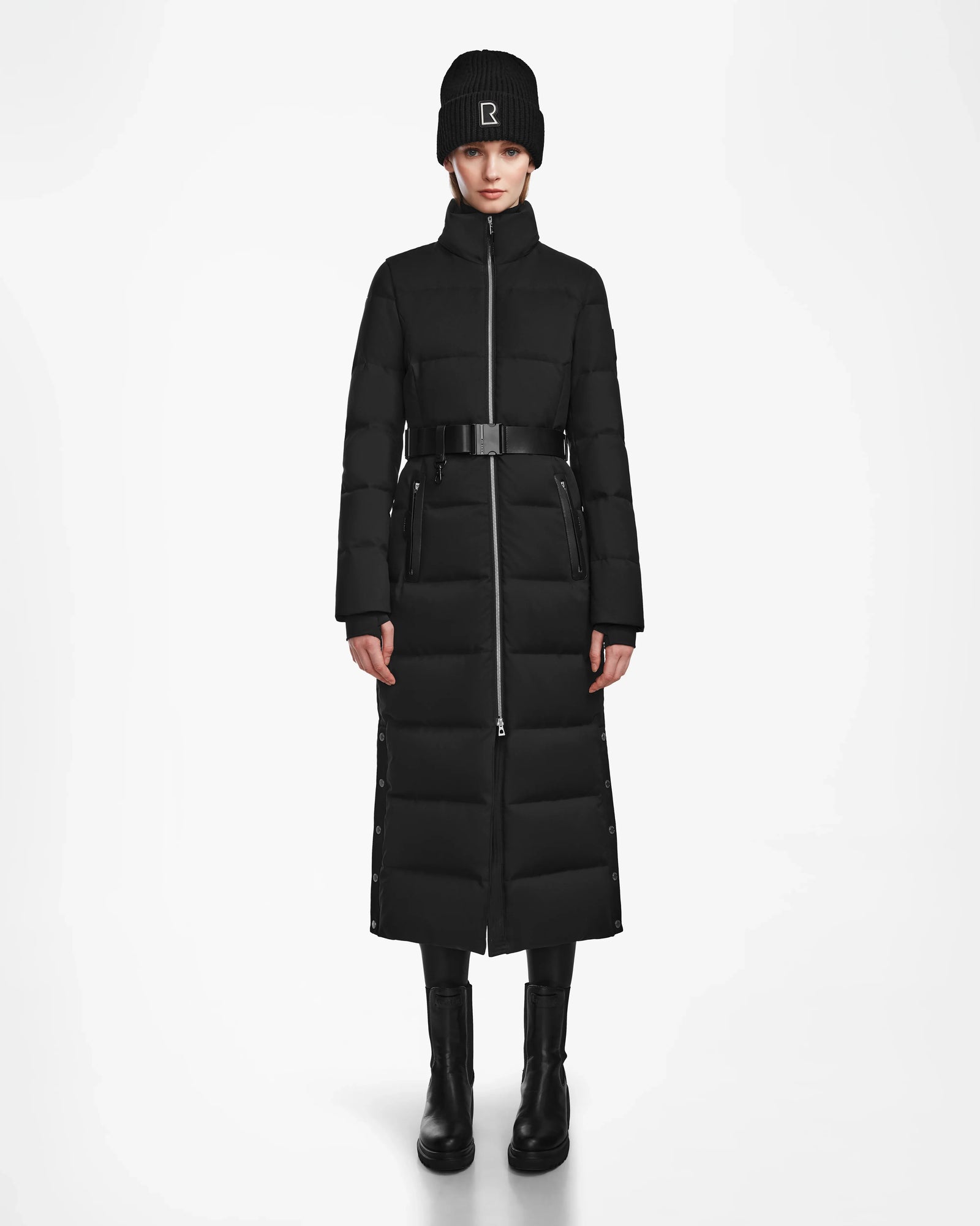 A person stands facing forward against a white background, wearing the Rudsak IZABELLA - L124541 Black women's long down quilted maxi coat, paired with a black beanie, leggings, and boots.