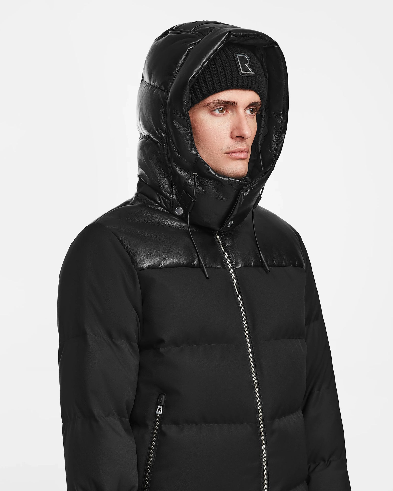 A person wears the Rudsak JACK - M124507 Black quilted jacket with hood and a black beanie, looking to the side against a plain white background.