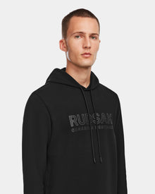 A man with short hair wears the Rudsak JARED - 6524533 Black hoodie featuring CANADIAN HERITAGE lettering, standing against a plain white background and looking forward with a neutral expression. This mens hoodie delivers effortless style.