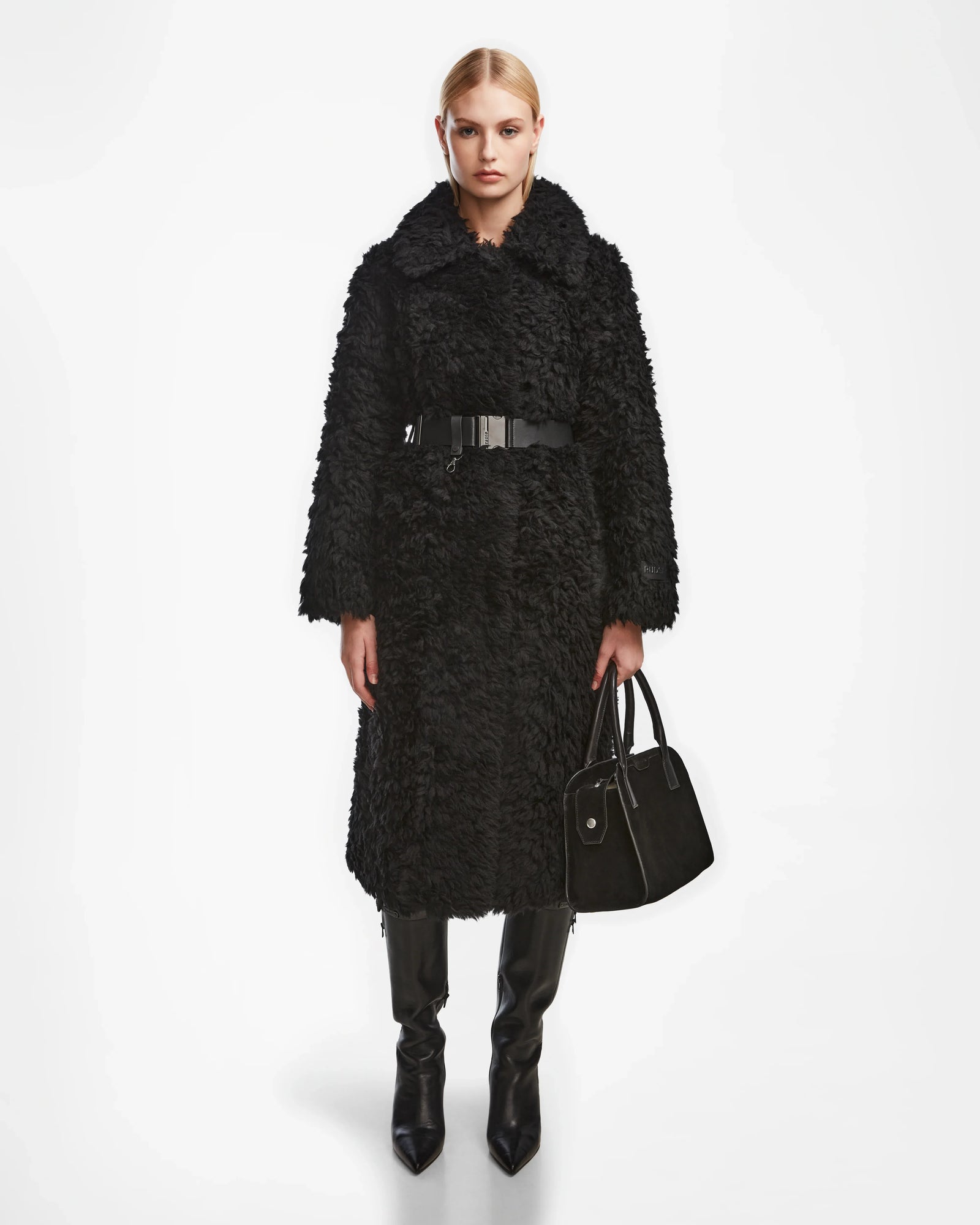 A woman stands in a studio wearing the Rudsak KAMILAH - 8124827 Black long faux fur coat with a wide collar and vegan leather belt, holding a black handbag, in black knee-high boots, her hair pulled back against a white backdrop.