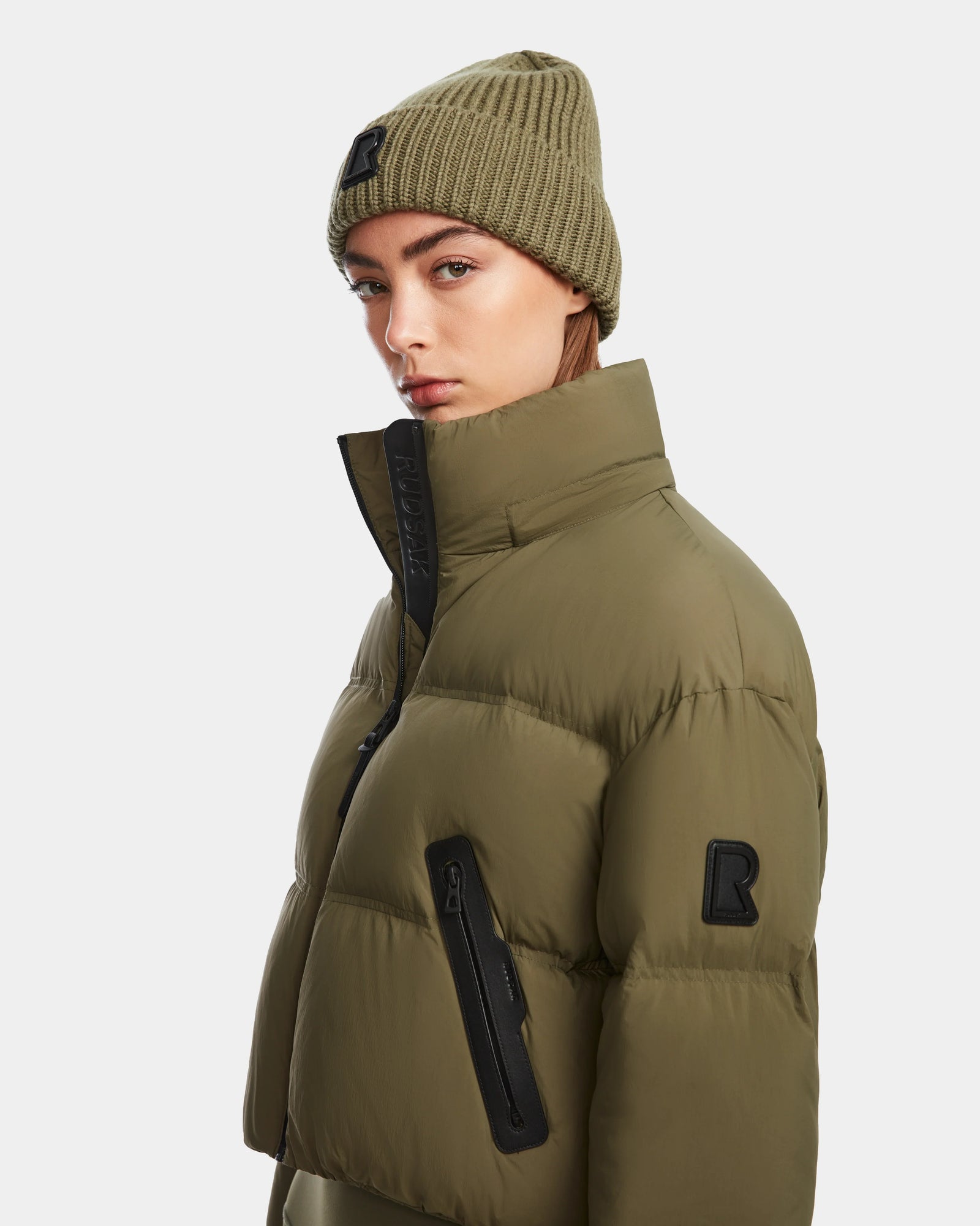 A woman wears the Rudsak KARLEE - 8124721 Loden Green cropped down puffer with black zippers and patches, paired with a matching knit beanie, looking at the camera against a plain white background.