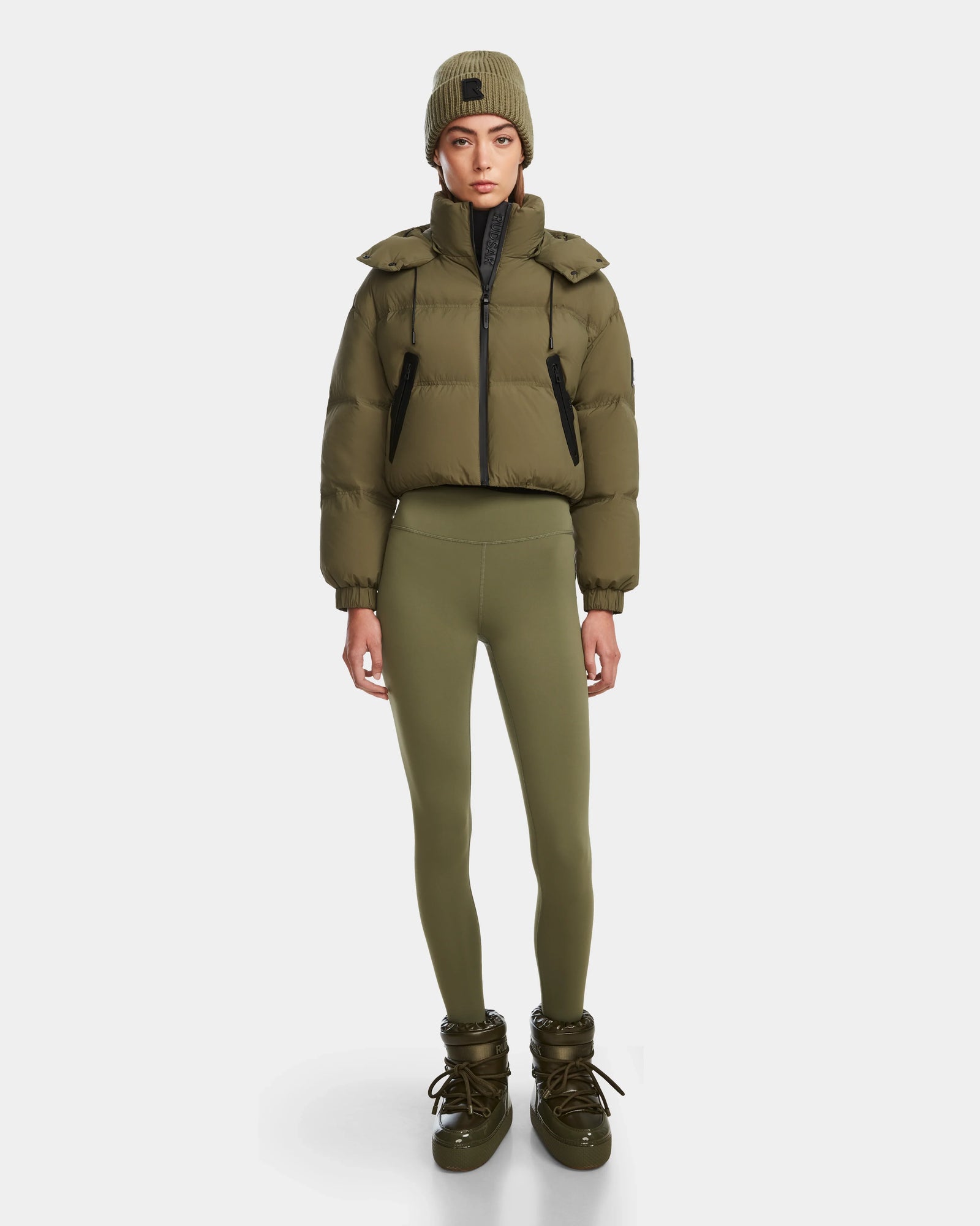 A woman faces forward in a Rudsak KARLEE - 8124721 Loden Green cropped down puffer jacket, matching leggings, knit beanie, and boots against a plain white background.