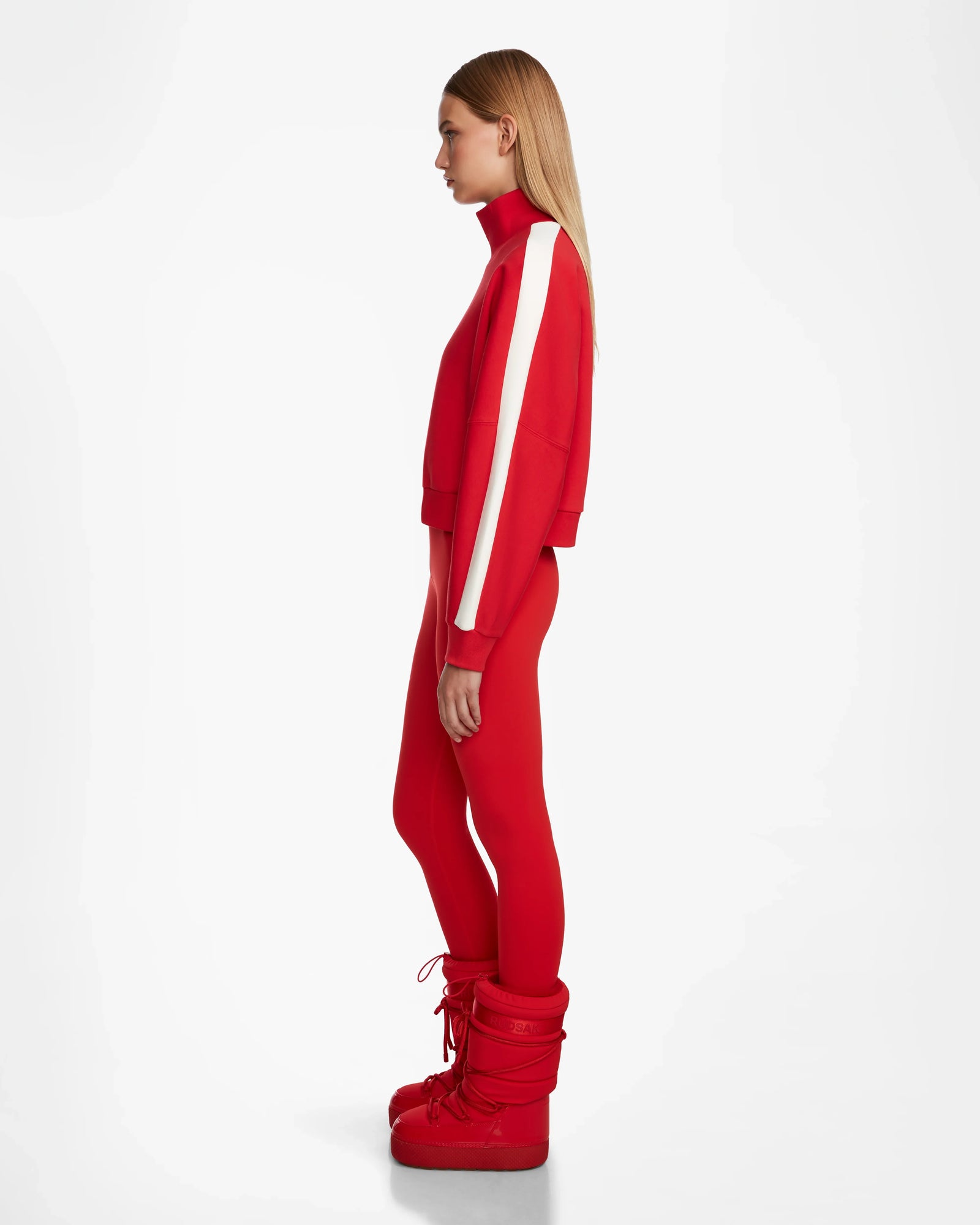 A woman stands in profile against a plain white background wearing the Rudsak KELLIE - 8524615 Flame Red top with white sleeve stripes and large red boots.