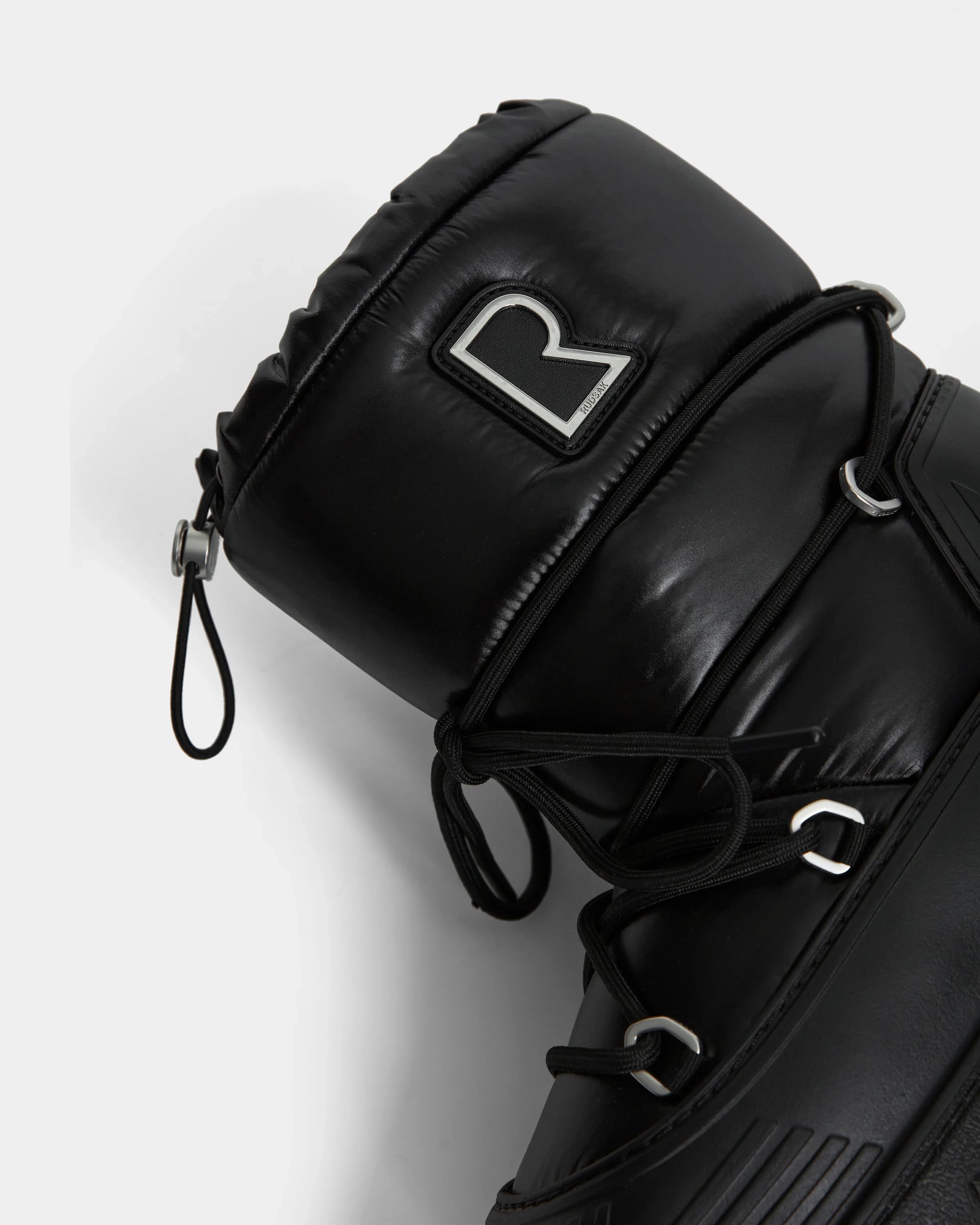 Close-up of the Rudsak KIKA - L224506 Black women’s winter boot featuring padded black material, black laces, silver eyelets, a bold metallic R logo on the upper cuff, and a chunky rubber sole, shown against a plain white background.