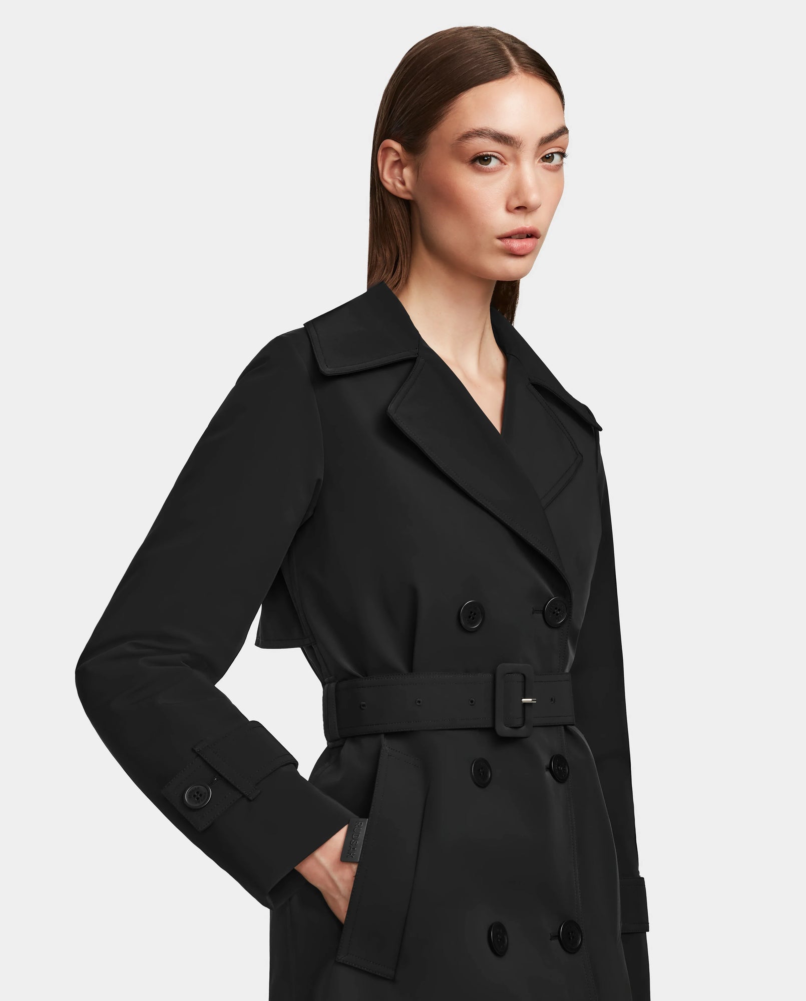 A woman with straight brown hair wears the Rudsak KIMBERLY - 8124801 Black women’s trench coat, featuring double-breasted buttons and a belt, standing against a plain light background.