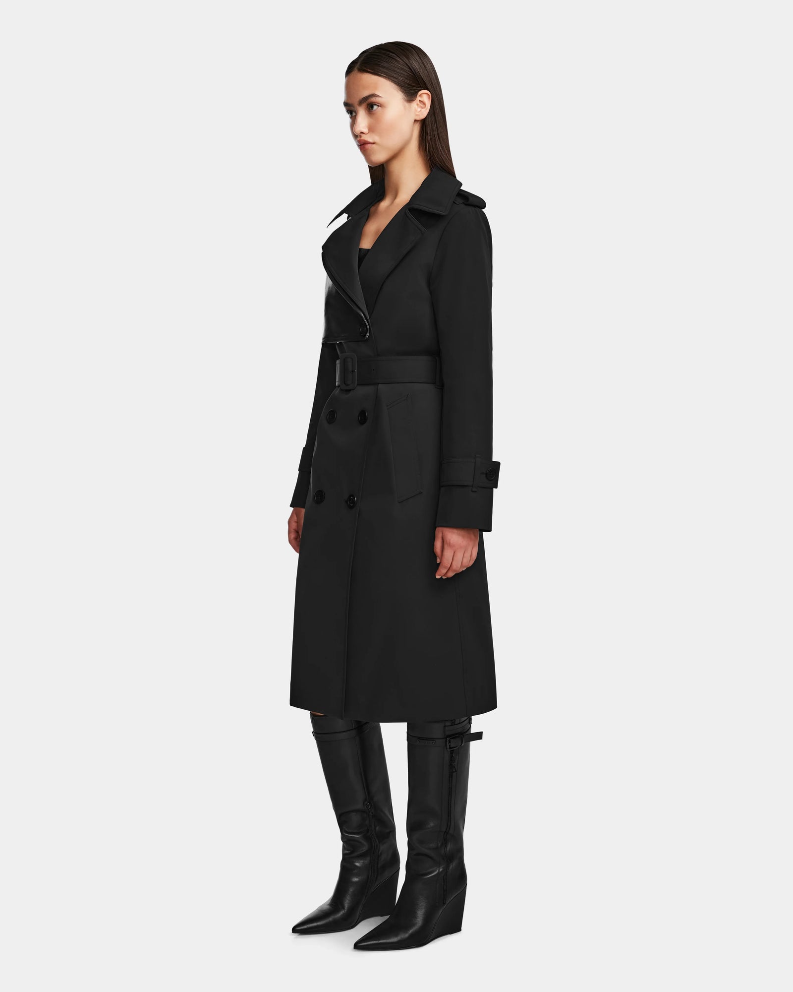 A woman in the Rudsak KINDA - 8124690 Black long trench coat with leather details and a belted waist, paired with black knee-high boots, stands against a plain light background, facing slightly to the right.