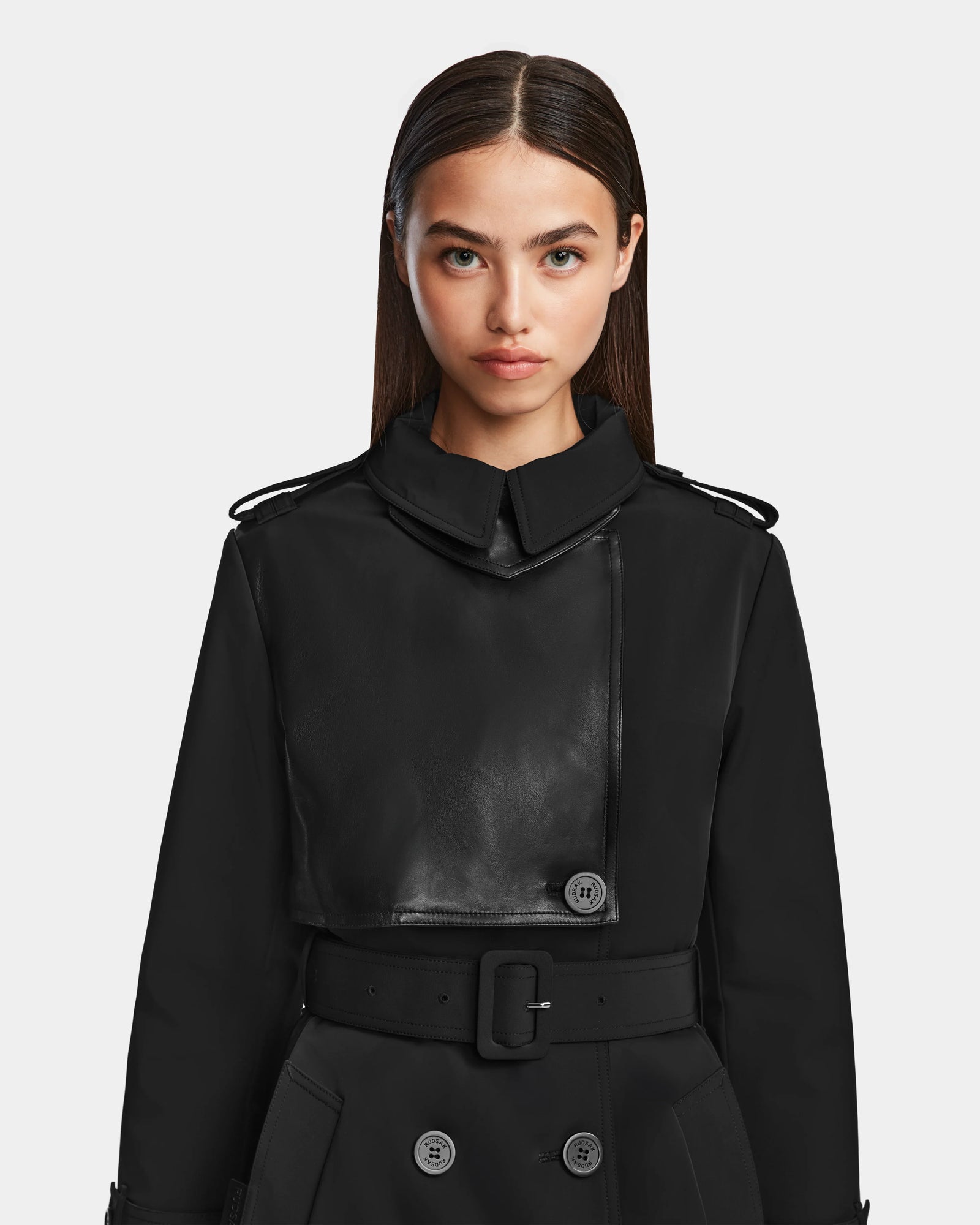 A woman with straight brown hair models the Rudsak KINDA - 8124690 Black leather trench coat with a belted waist, standing against a light gray background and looking directly at the camera with a neutral expression.