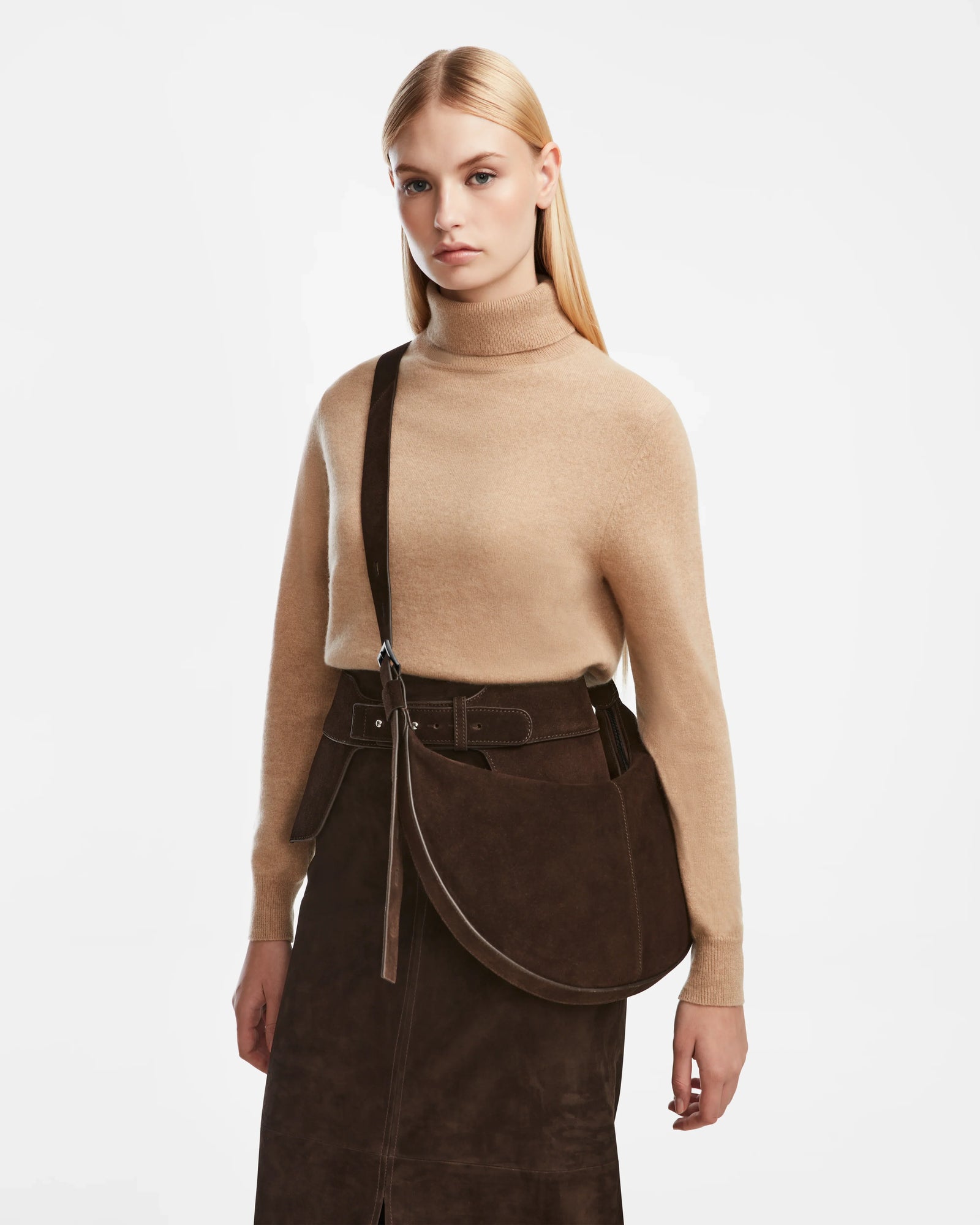 A woman with long blonde hair stands against a white background, wearing a beige turtleneck, dark brown suede skirt, and carrying the Rudsak LANISSA S - 8324520 Espresso suede hobo bag.