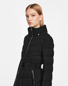 A woman with straight blonde hair models the Rudsak MALAYA M - L123554 Black women’s down puffer jacket, featuring a belted waist, herringbone quilting, and leather accents, made from recycled matte shell, against a plain white background.