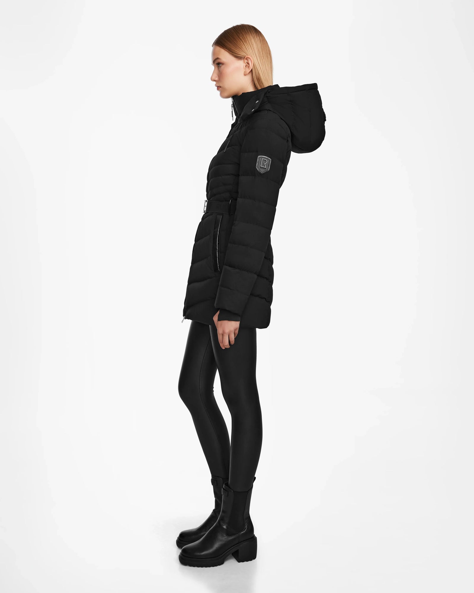 A woman in profile wears the Rudsak MALAYA M - L123554 Black womens down puffer jacket with a herringbone quilt, paired with black leggings and ankle boots, against a plain white background.