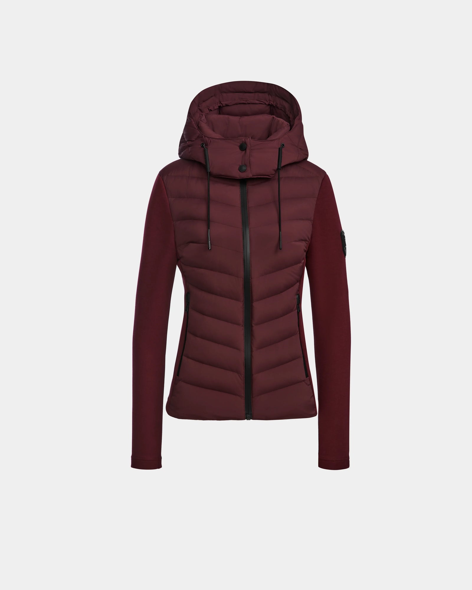 The Rudsak MALYA M - L123547 Wine is a womens burgundy down quilted jacket featuring a front zipper, black drawstrings, quilted body, smooth long sleeves, fitted silhouette, and a small black patch on the upper left sleeve.