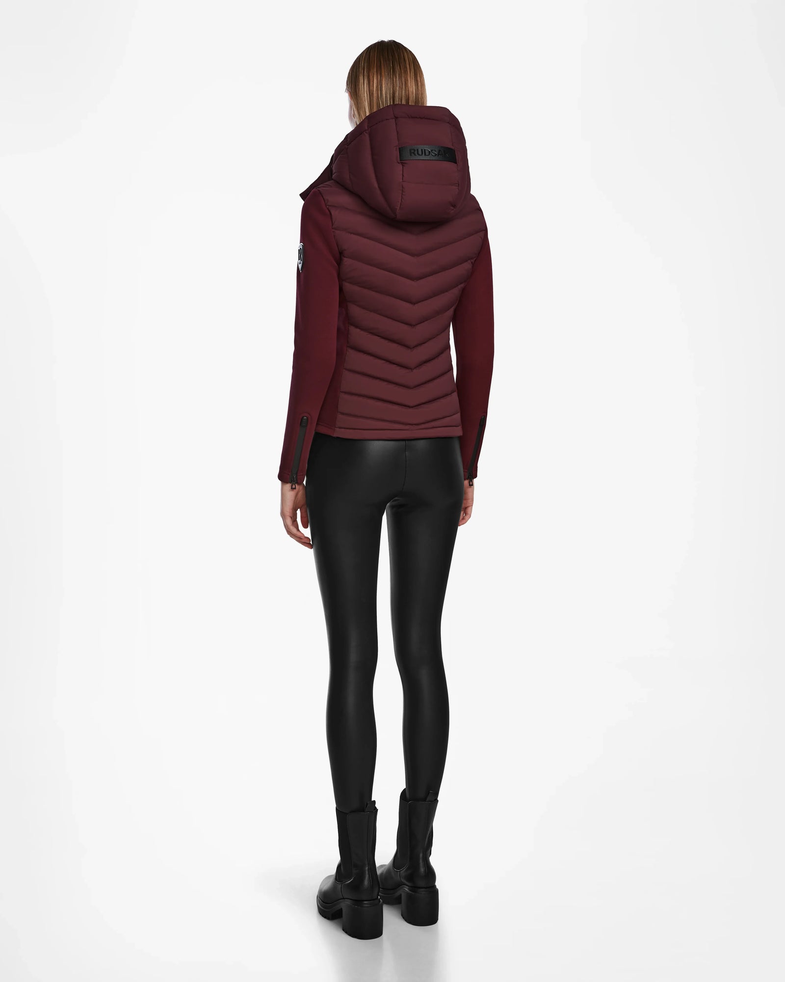A person stands facing away, wearing the Rudsak MALYA M - L123547 Wine down quilted jacket with a hood, paired with black shiny leggings and black boots against a plain white background.