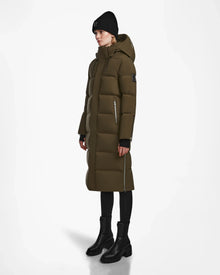 A person wearing the Rudsak MAO M - 8123598 Army women’s down puffer coat with a hood, black beanie, leggings, and ankle boots stands against a plain white background.