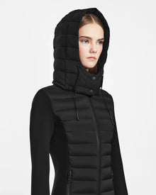A woman wears the Rudsak MARRY M - L123548 Black women’s puffer jacket with the hood up, looking serious and facing slightly right against a plain white background.