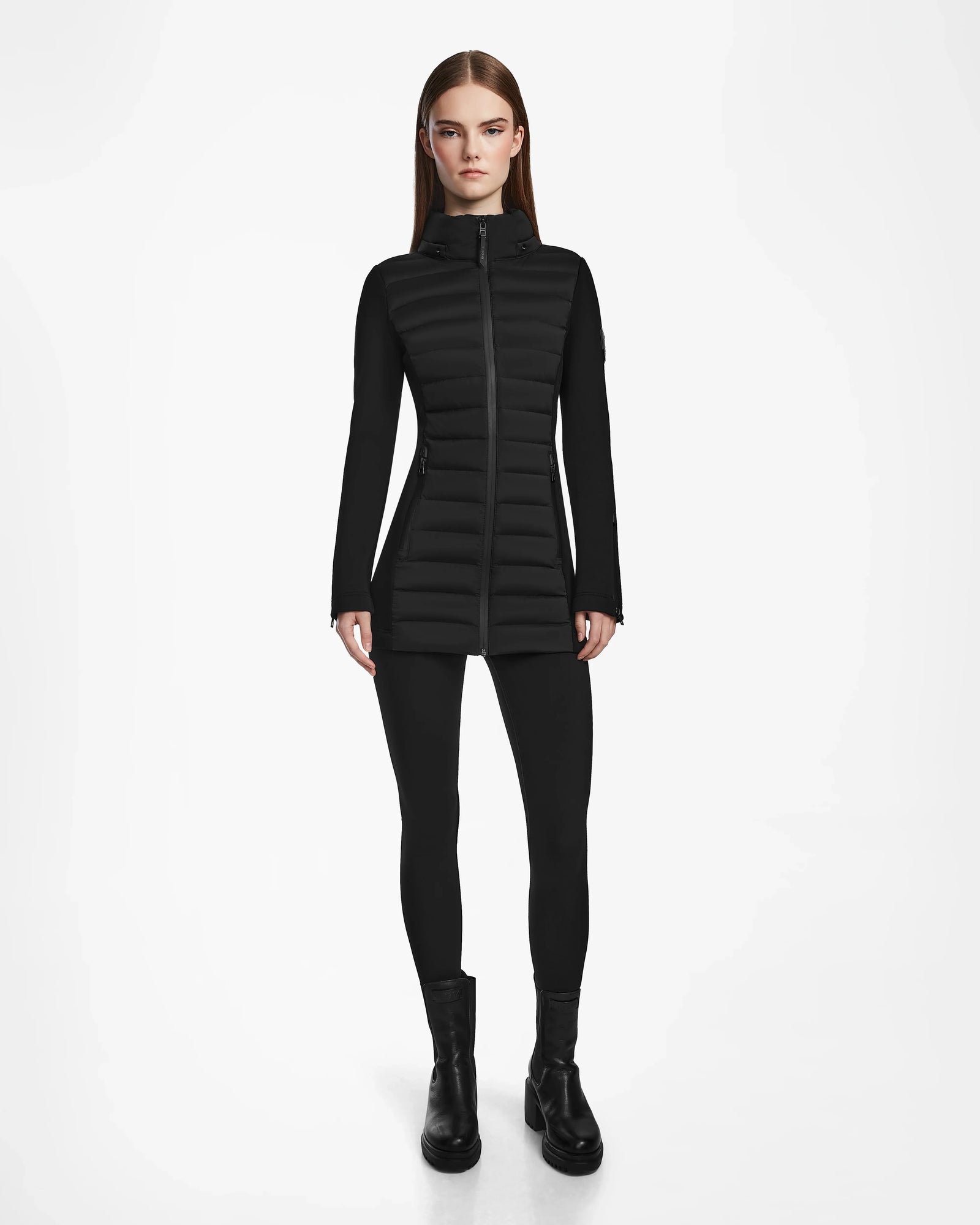 A woman stands facing forward, wearing the Rudsak MARRY M - L123548 Black women’s puffer jacket with a removable hood, paired with black leggings and boots against a plain white background.