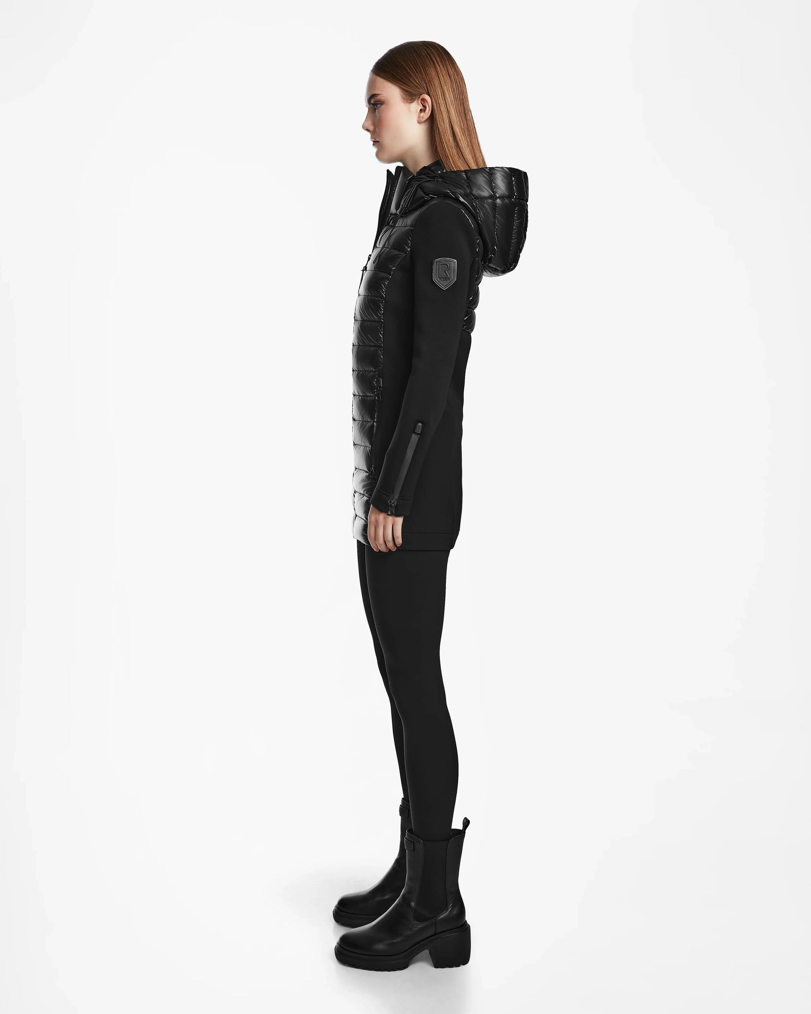 A woman with straight light brown hair stands in profile against a white background, wearing the Rudsak MARRY - L121003 BLACK recycled down women’s puffer jacket, paired with black leggings and ankle boots.