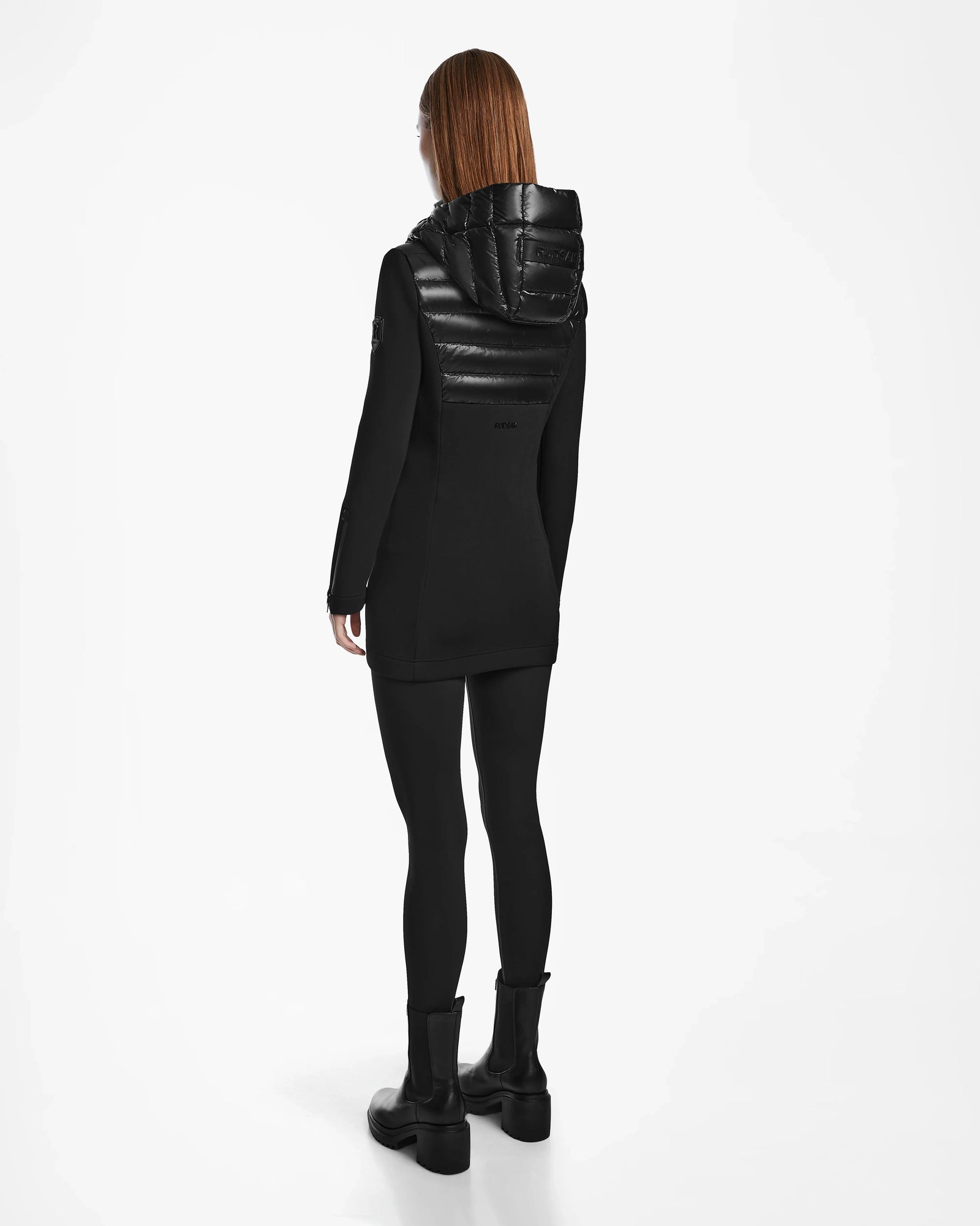 A person with long, straight auburn hair stands facing away, wearing the Rudsak MARRY - L121003 BLACK women’s puffer jacket, black leggings, and black boots against a white background.