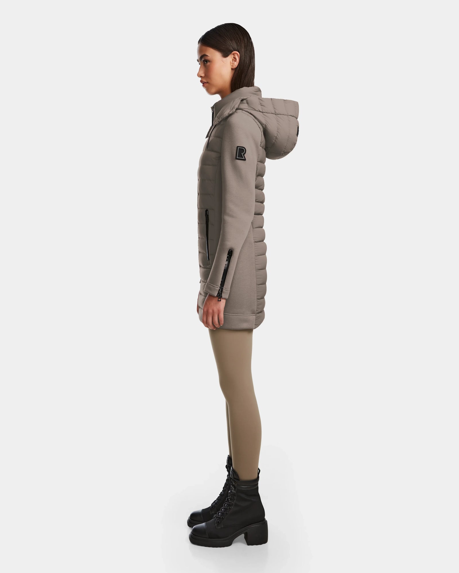 A woman in profile wears the Rudsak MERINA M - 8124030 Cement, a taupe quilted down jacket with a hood, paired with beige leggings and black lace-up boots, standing against a plain white background.
