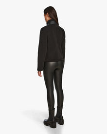 A person faces away, wearing the Rudsak MILANA - 8524604 Black womens teddy sweater with vegan leather details, paired with shiny leggings and black ankle boots, against a plain white background.