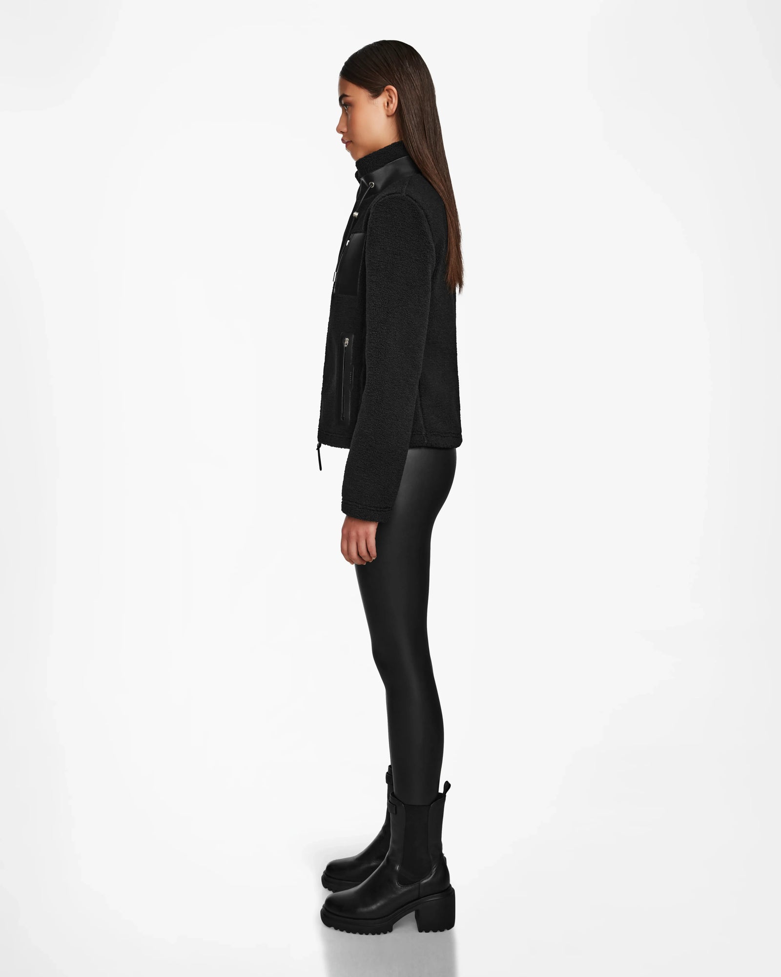 A young woman stands in profile against a white background, wearing the Rudsak MILANA - 8524604 Black zip-up jacket with vegan leather details, fitted black pants, and chunky black boots. Her straight hair is left loose.