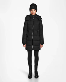 A person stands facing forward wearing Rudsak’s MIRIAM - L122523 BLACK women’s down puffer coat, styled with a black knit beanie, black leggings, and black ankle boots against a plain white background.
