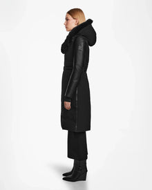 A woman stands in profile against a plain white background wearing the Rudsak MISALA - L124512 Black womens long down coat with a fur-trimmed hood, paired with black pants and black heeled boots.