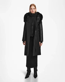 A woman faces forward wearing the Rudsak MISALA - L124512 Black long down coat with a fur-trimmed hood, paired with a black skirt, high-neck inner layer, and black heeled boots against a plain white background.