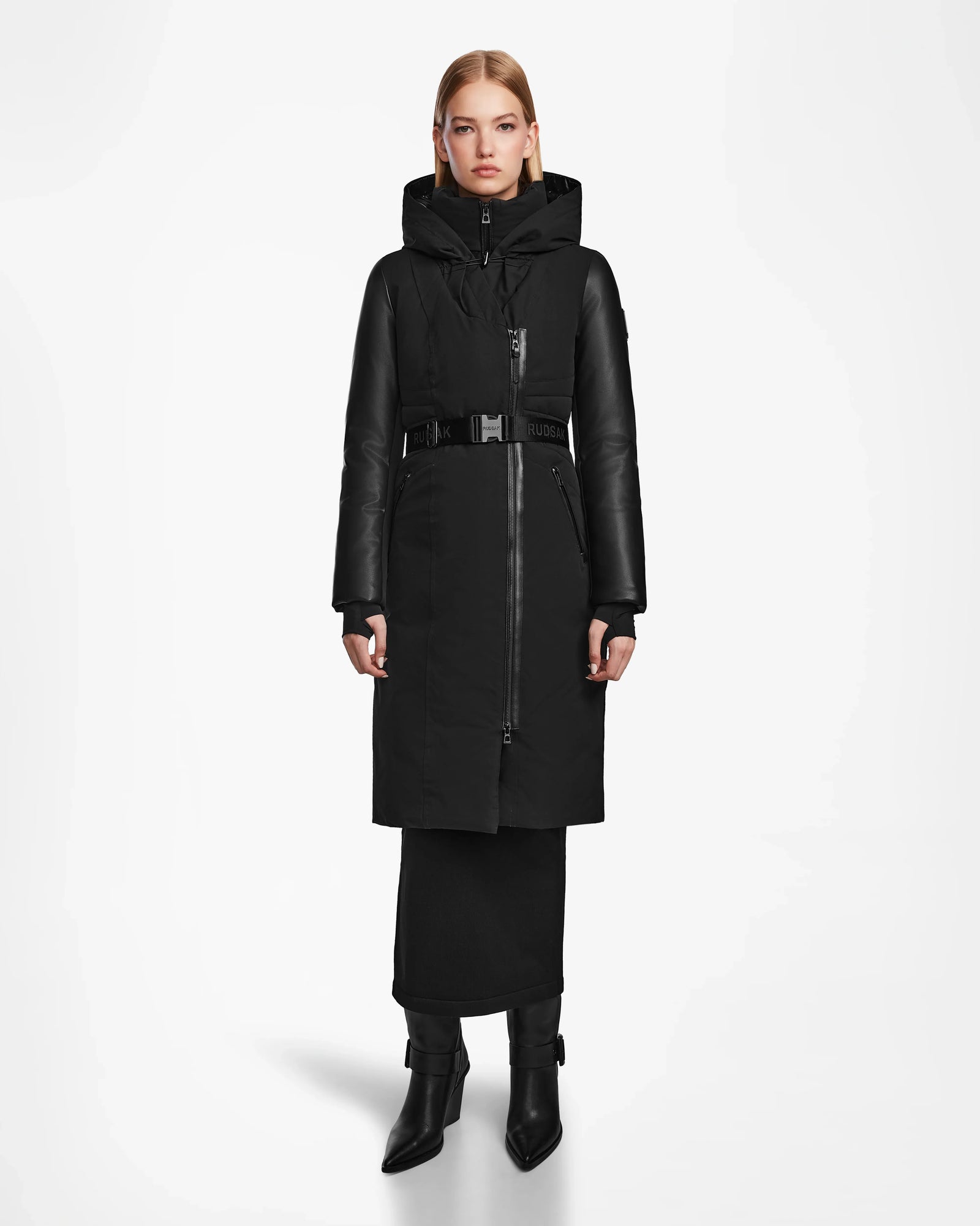 A woman faces forward in the Rudsak MISALA - L124512 Black water-resistant winter coat with a high collar and hood, paired with a black skirt and heeled ankle boots, against a plain white background.