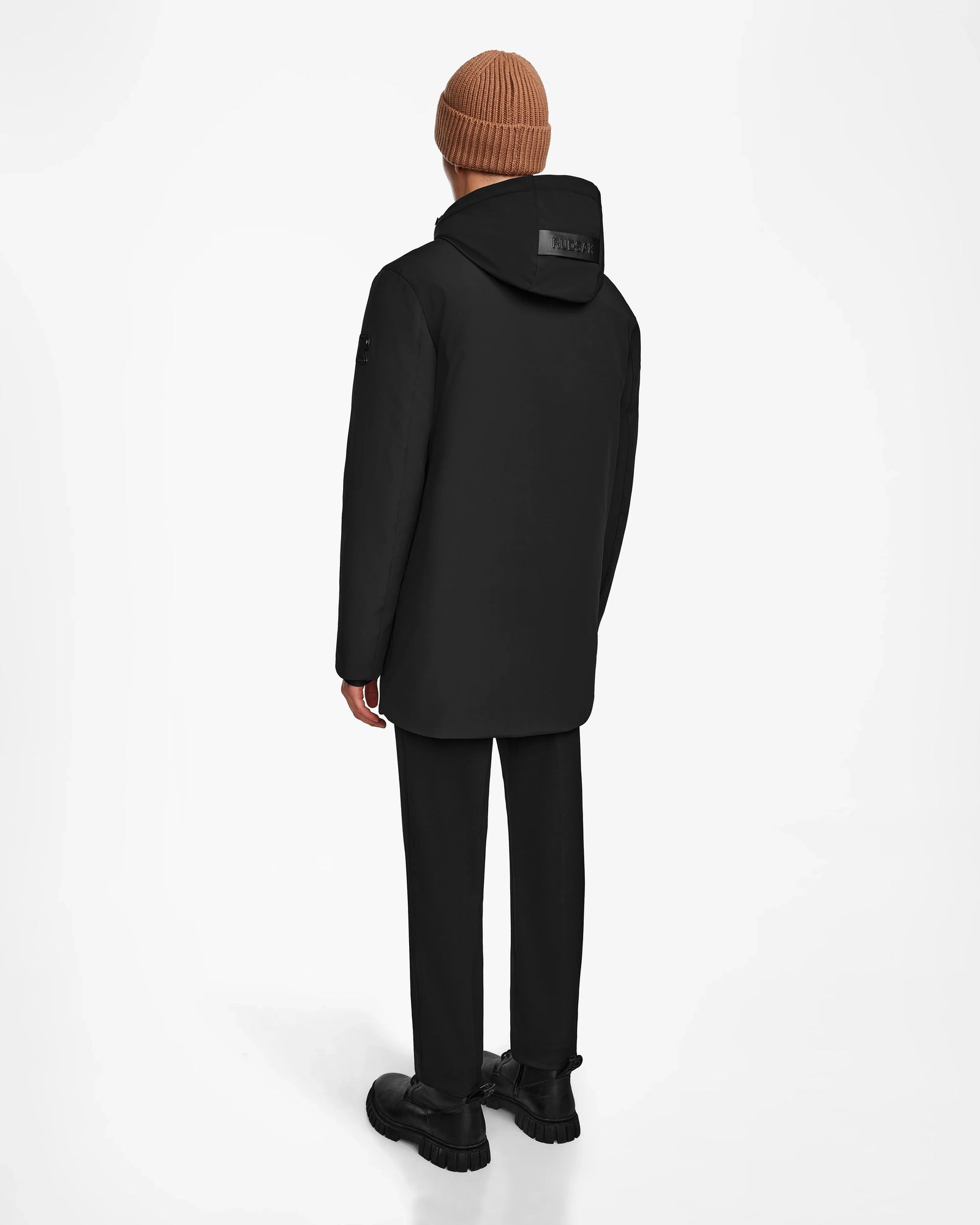 A person wearing the Rudsak NATAN - M124519 Black men’s parka stands facing away against a plain white background, paired with black pants, black shoes, and a brown knit beanie.