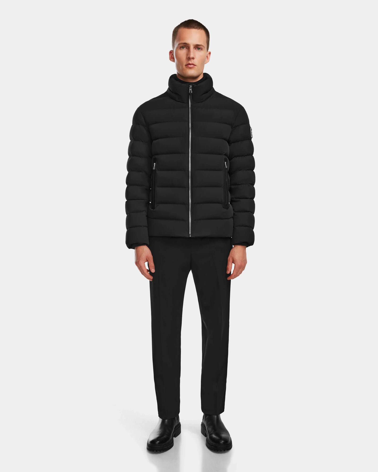 A man faces forward wearing the Rudsak NAVAN - 6124551 Black mens quilted down puffer with wind and water-repellent material, paired with black pants and shoes, against a plain white background.