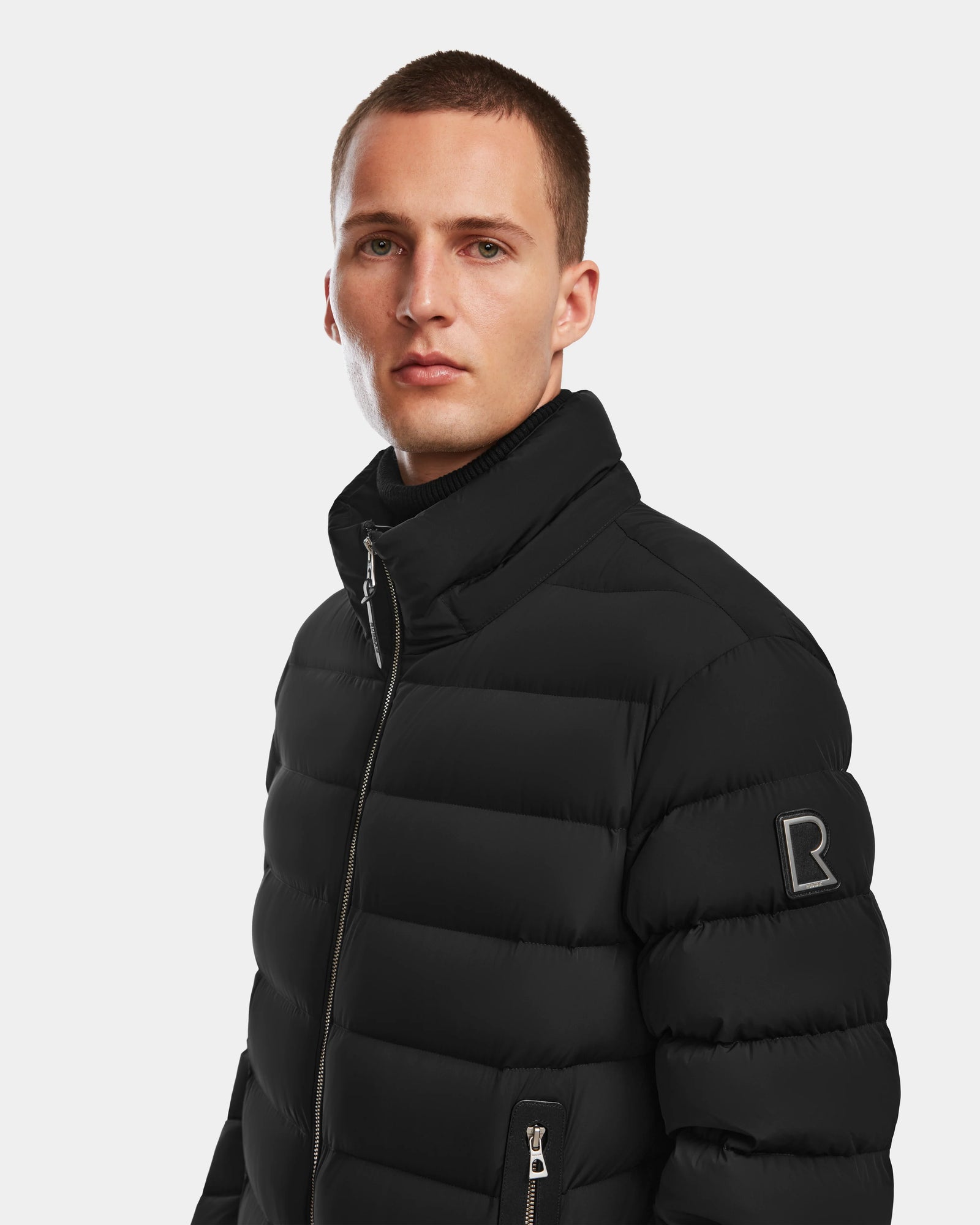 A young man with short hair wears the Rudsak NAVAN - 6124551 Black mens quilted down puffer jacket, featuring a visible zipper and R logo patch on the sleeve, standing against a plain light background.