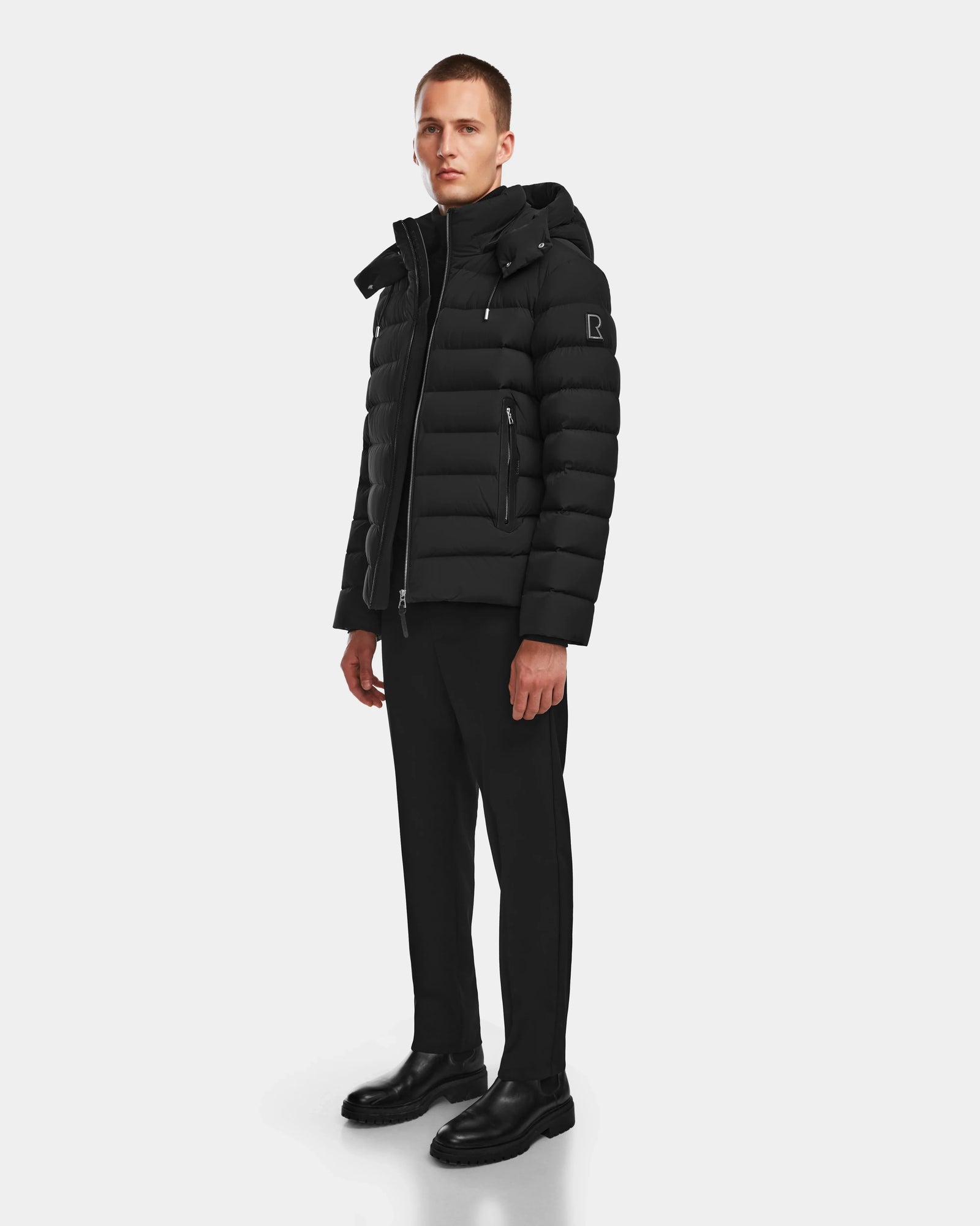 A man stands against a plain background wearing the Rudsak NAVAN - 6124551 Black mens quilted down puffer jacket with RDS-certified down, paired with a black turtleneck, pants, and shoes. He faces slightly to the side with a neutral expression.