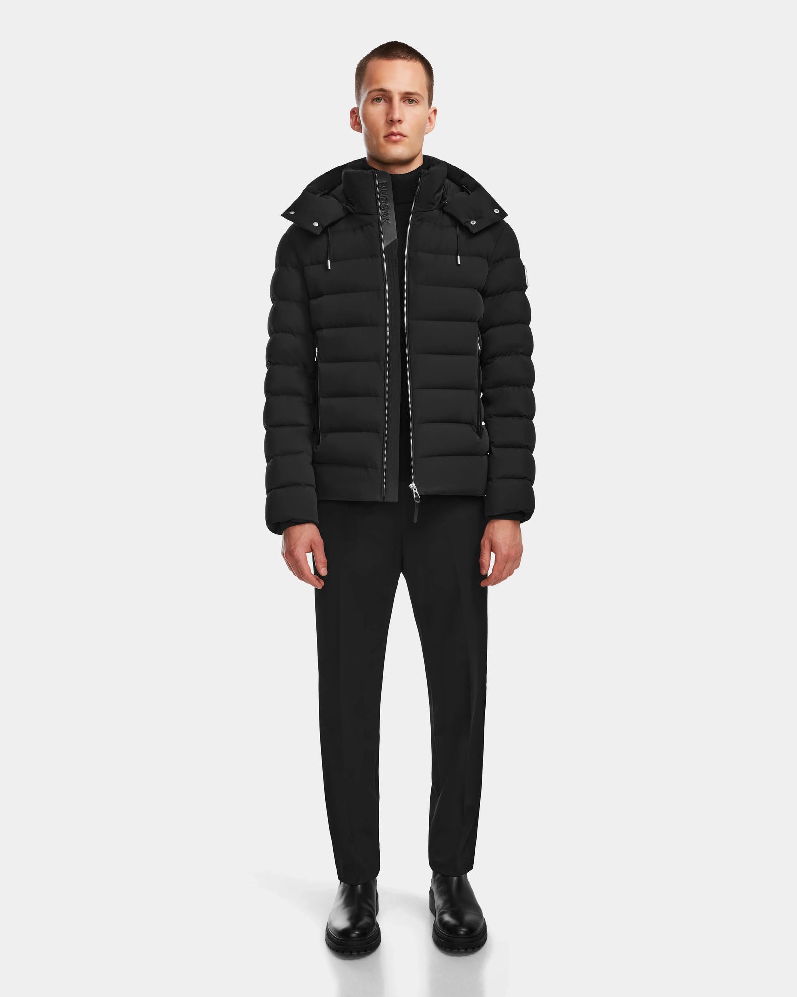 A person stands facing forward wearing the Rudsak NAVAN - 6124551 Black mens quilted down puffer jacket with a hood, paired with black pants, black shoes, and a black shirt against a plain white background.