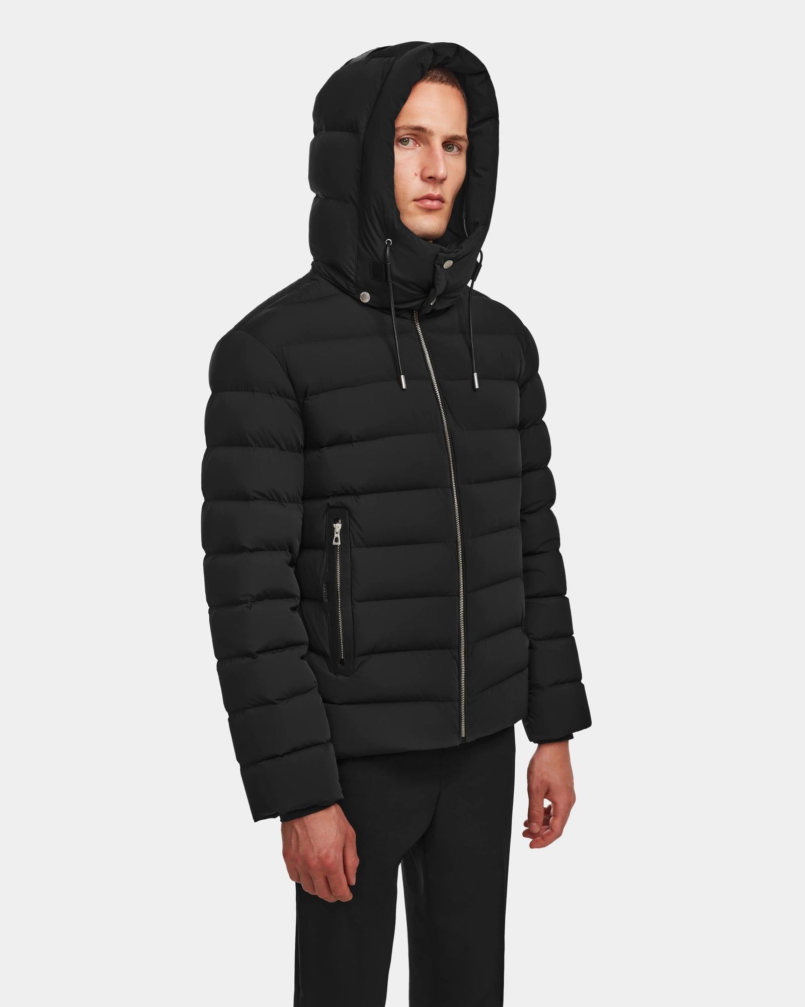 A person wears the Rudsak NAVAN - 6124551 Black men’s quilted down puffer jacket, featuring vertical quilting, zippered side pockets, RDS-certified down, and a wind- and water-repellent finish, against a plain white background.