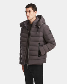 A person wearing the Rudsak NAVAN - 6124551 Grey mens down puffer jacket with a hood stands with one hand in their pocket against a plain light background.