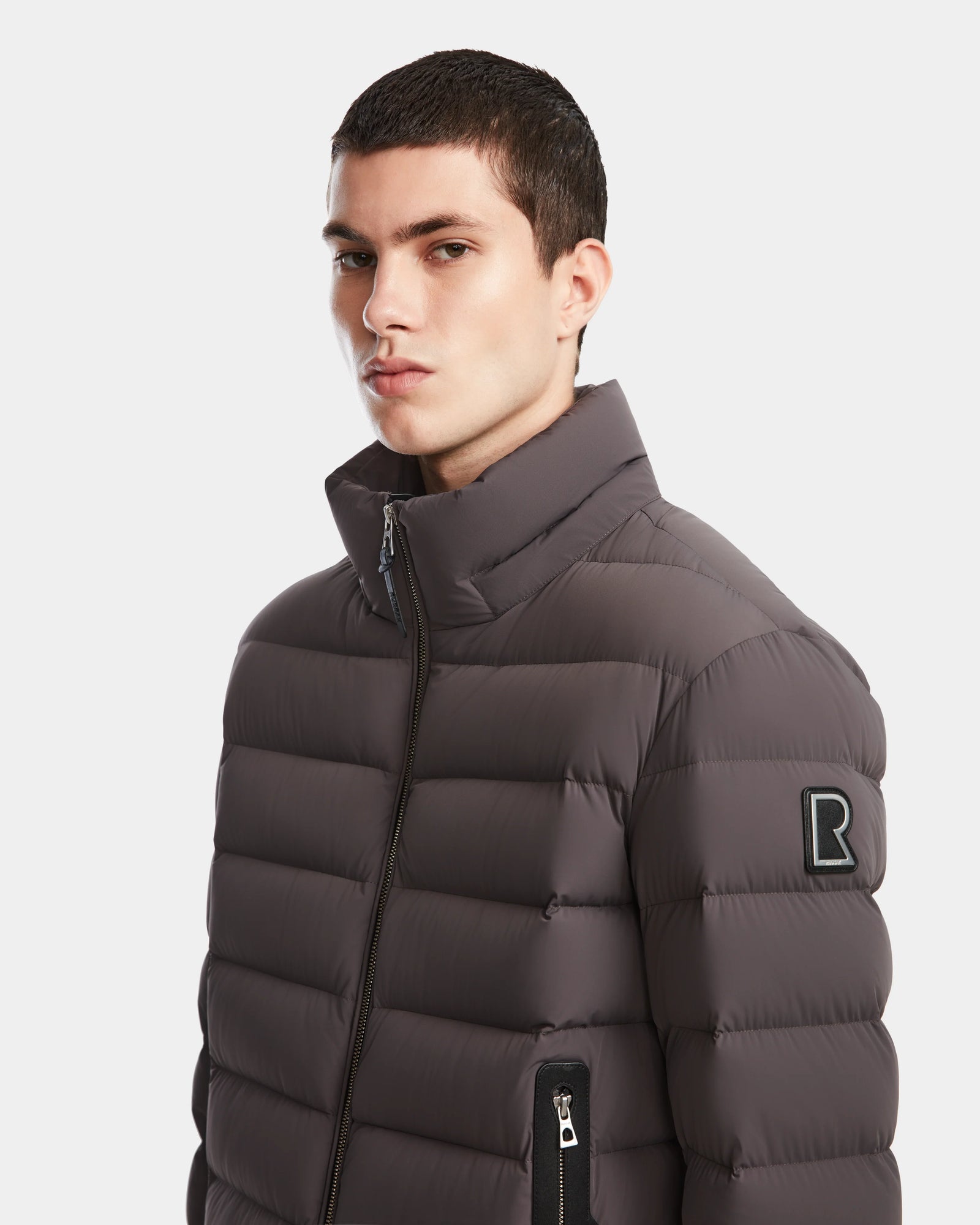 A young man with short dark hair wears the Rudsak NAVAN - 6124551 Grey mens quilted down puffer jacket with a high collar and a black R patch on the sleeve, looking at the camera against a plain white background.