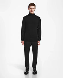 A person stands facing forward against a plain white background, wearing the Rudsak NICOLAS - 6524528 Black mens cashmere turtleneck sweater, paired with black pants and black boots.