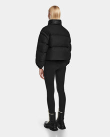 A person with long blonde hair is standing and facing away, wearing the Rudsak OPRAH - 8124708 Black womens cropped down puffer jacket, black leggings, and black boots with white fur trim against a plain light background.
