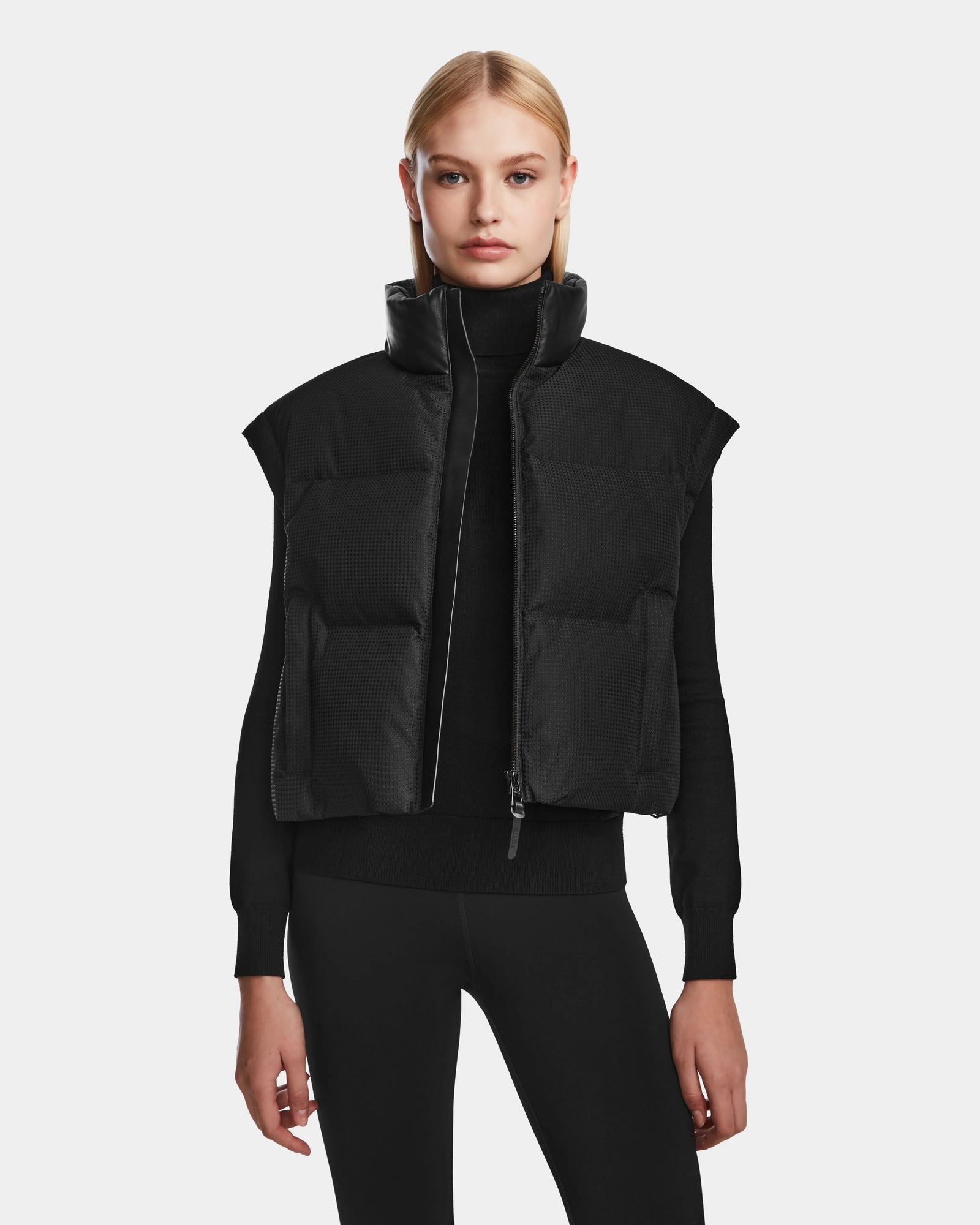 A woman with straight blonde hair models the Rudsak OPRAH - 8124708 Black women’s cropped down puffer vest over a black long-sleeve top and pants, standing against a plain white background.