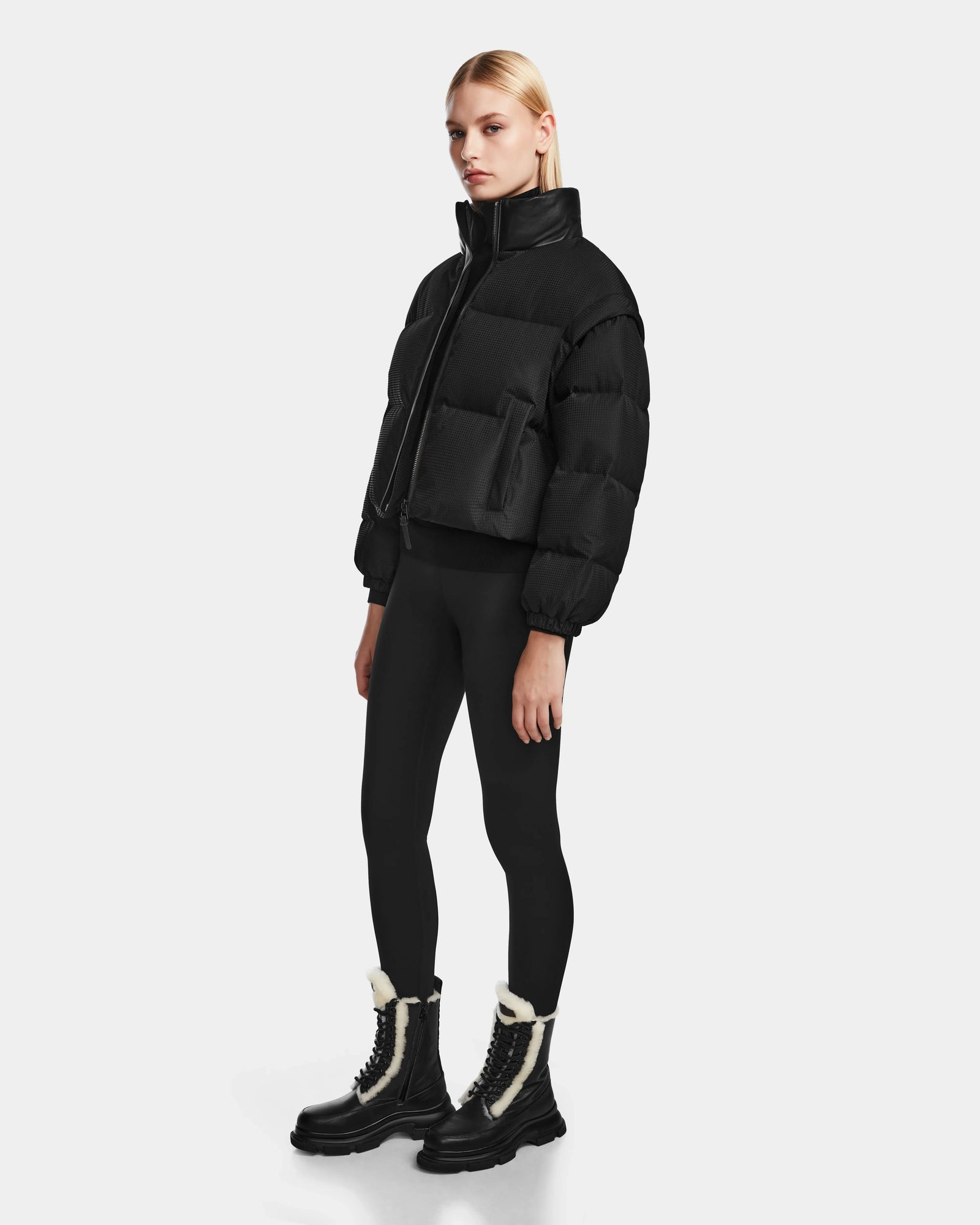 A woman with straight blonde hair and a neutral expression wears the Rudsak OPRAH - 8124708 Black women’s cropped down puffer, paired with black leggings and black fur-trimmed lace-up boots, against a plain white background.